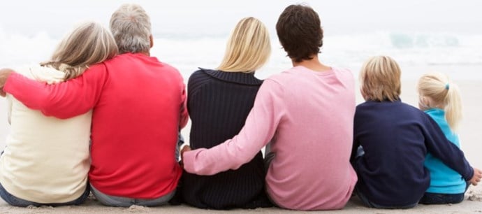 Tre generationers familj ses bakifrån sittande tillsammans på en strand.